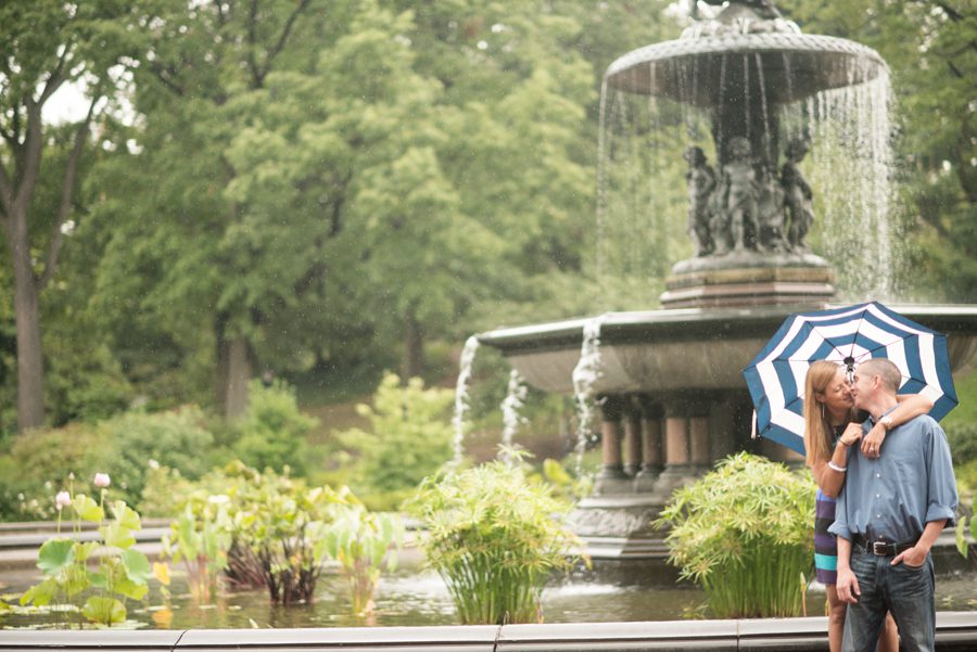 Erin and Tim pose with an umbrella by a fountain during their rainy engagement session in Central Park with Ben Lau Photography.