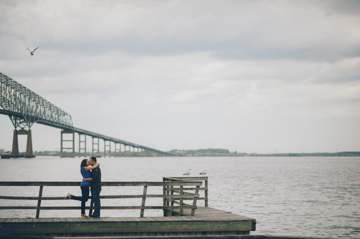 Michelle and Bryan embrace on a dock with the Key Bridge in the background during their Baltimore engagement session with Ben Lau Photography.