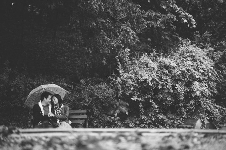 A couple sits on a bench with an umbrella during their rainy engagement session in Prospect Park with NYC wedding photographer Ben Lau.