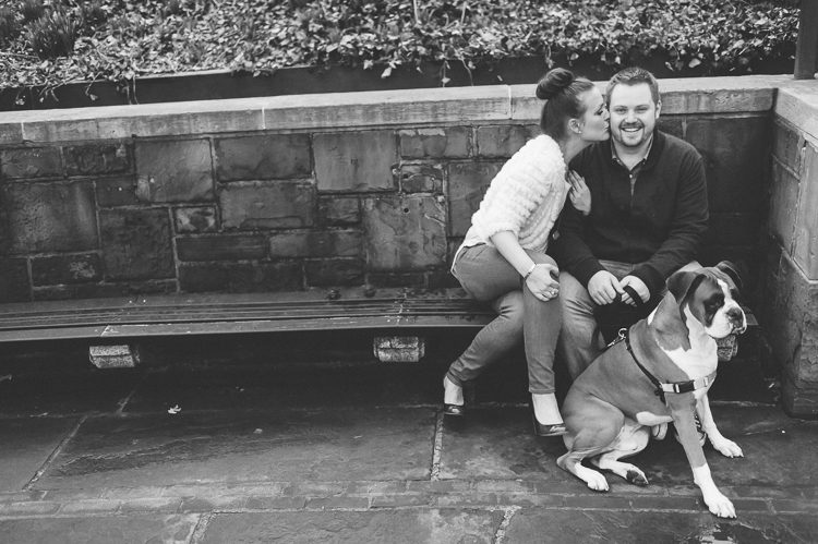 Girl kisses guy while sitting on a bench with their dog, during their engagement session in Central Park. Captured by NYC wedding photographer Ben Lau.