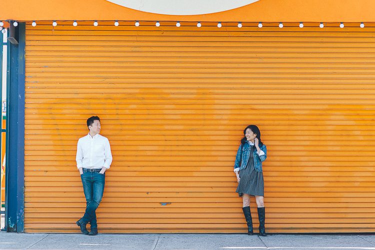 Couple pose against an orange gate during their engagement session at Coney Island. Captured by NYC wedding photographer Ben Lau.