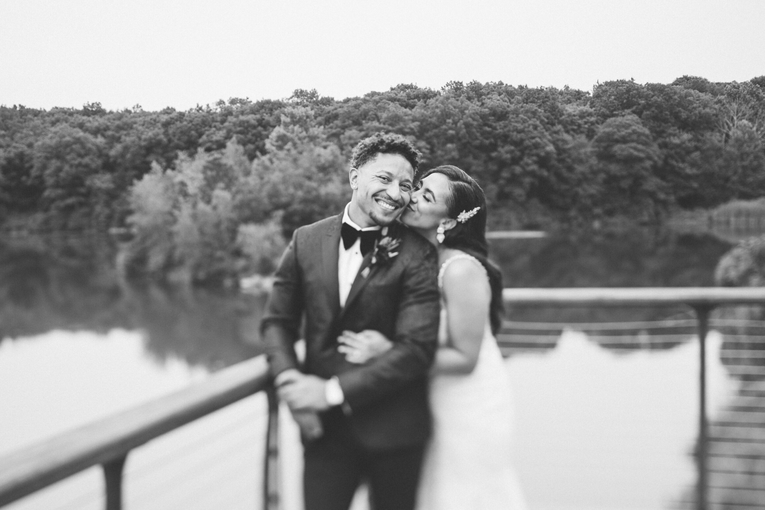 Black and white portrait of Alliee and Brandon embracing on the dock by the lake.